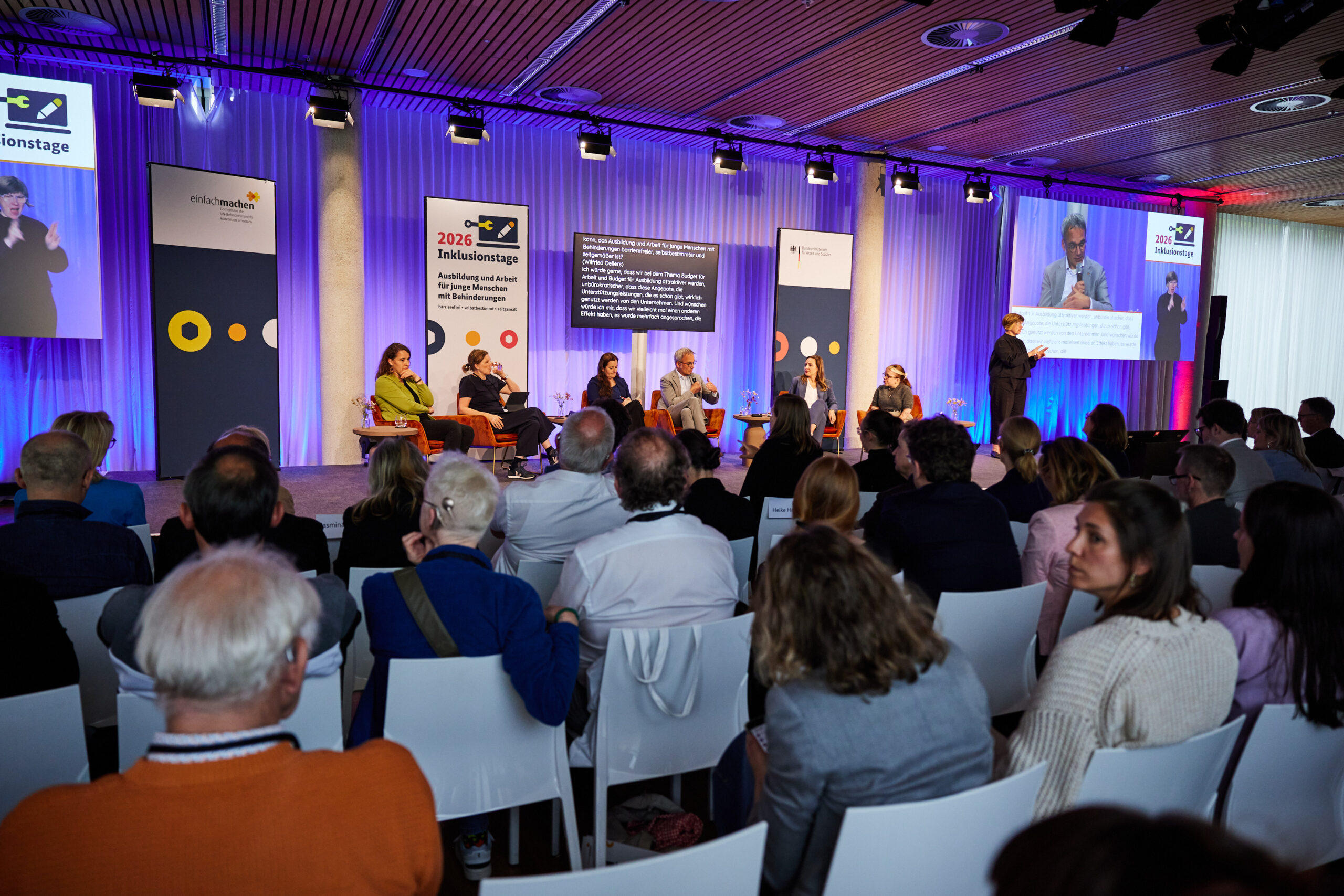 Podiumsdiskussion bei den Inklusionstagen 2026 mit Publikum, Gebärdensprachdolmetscherin, Live-Untertiteln und Übertragung auf zwei Leinwänden.