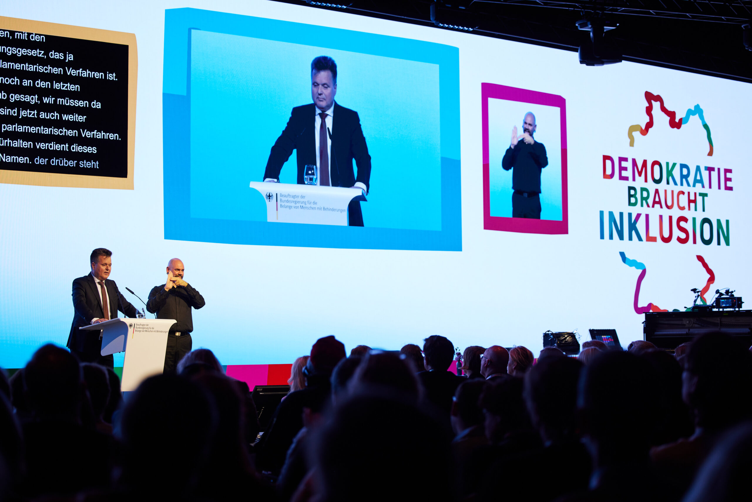 Jürgen Dusel, Beauftragter der Bundesregierung für die Belange von Menschen mit Behinderungen, spricht am Rednerpult. Ein Gebärdensprachdolmetscher übersetzt die Rede, die mit Live-Untertiteln auf einer großen Leinwand übertragen wird.